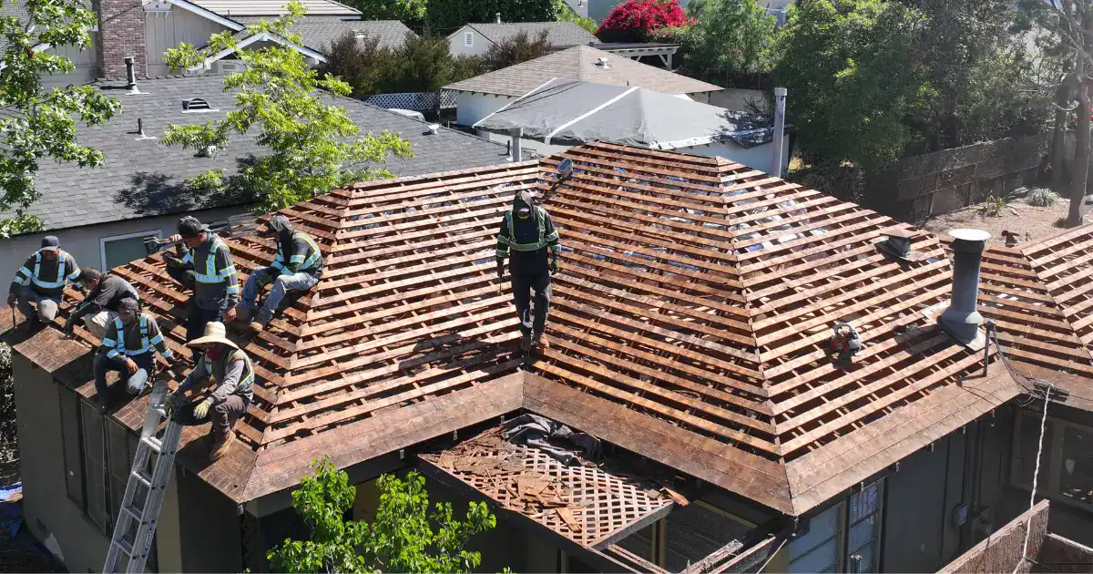 Aerial view of a residential roof replacement project in Southern California showing a roofing crew performing a complete tear-off. Workers are removing old roofing materials and exposing the wood battens and roof deck on a multi-faceted hip roof home surrounded by neighboring houses and mature trees.