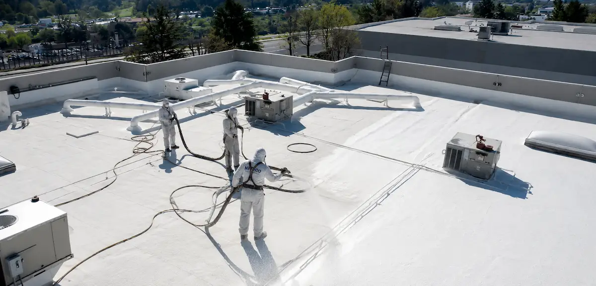 Method Roofing Group crew applying spray polyurethane foam (SPF) topcoat on a commercial flat roof with rooftop HVAC equipment, ductwork, and parapet walls visible